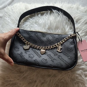 Juicy Couture svart axelväska med kedja - Svart axelväska från Juicy Couture i syntetmaterial med präglad logga över hela väskan. Guldig kedja med hängande berlocker i form av hjärta, krona och JC-logga. Dragkedja upptill och innerfoder med tryckt logomönster. Perfekt för dig som gillar statement-accessoarer.