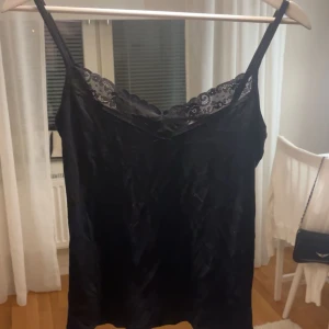 Svart linne med spets  - Supersnygg svart linnetopp i sammet från 157 med tunna axelband och spetsdetalj längs v-ringningen. Perfekt att styla med jeans eller kjol för en trendig look. Mjuk och glansig yta som ger en lyxig känsla.
