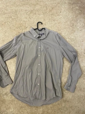 Grå skjorta från Tommy Hilfiger - Klassisk grå skjorta från Tommy Hilfiger med button down-krage och små loggan broderad på bröstet. Skjortan har långa ärmar, vita knappar och är tillverkad i bomull. Perfekt för dig som gillar stilrena och enkla plagg.