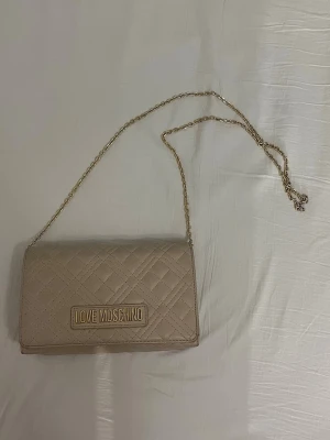  Love Moschino axelväska  - Säljer en beige axelväska från Love Moschino med guldfärgad kedjerem och logga framtill. Väskan har ett elegant rutmönster och stängs med magnetknapp. Perfekt storlek för det viktigaste och ger en lyxig touch till din outfit.