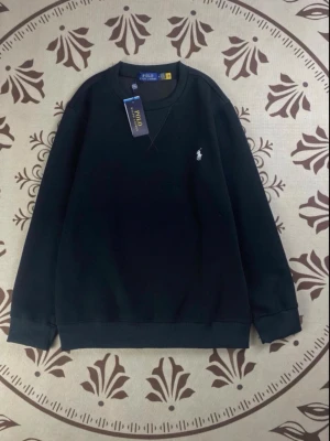 Svart sweatshirt från Polo Ralph Lauren - Klassisk svart sweatshirt från Polo Ralph Lauren med rund halsringning och den ikoniska vita logon broderad på bröstet. Tröjan har ribbade muddar vid ärmslut och nederkant, perfekt för en clean och stilren look.
