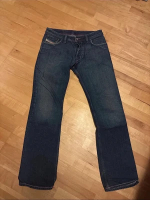 Diesel jeans mörkblå  herr 31/32 - Säljer ett par klassiska blå jeans från Diesel, tillverkade i Italien. Jeansen har raka ben, fem fickor och snygga detaljer på bakfickorna. Materialet är 100% bomull och färgen är mörkblå med lätt slitning. Perfekta till en avslappnad stil.