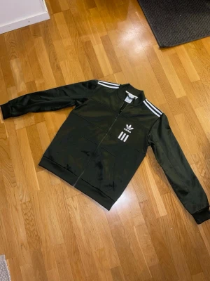 Grön Adidas vindjacka med vita ränder - Säljer en svart Adidas vindjacka med klassiska vita ränder på axlarna och logga både fram och bak. Jackan har dragkedja hela vägen, två fickor och ribbade muddar. Tillverkad i polyester och har en sportig, clean look som funkar till allt.