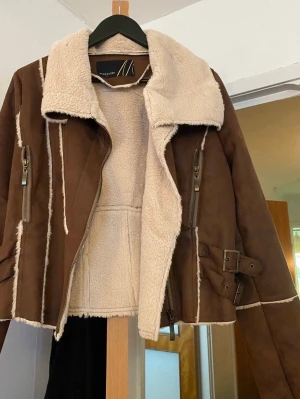 Brun teddyjacka  - Snygg brun teddyjacka med krage och detaljer i beige fuskpäls. Jackan har dragkedjor på sidorna, spännen vid midjan och markerade sömmar. Perfekt för dig som vill ha en cool och varm look under hösten och vintern.