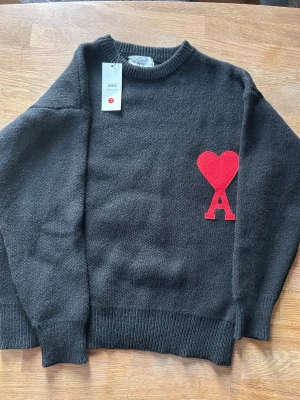 Svart stickad tröja från Ami paris  - Svart stickad tröja från Ami Alexandre Mattiussi med röd hjärt- och A-detalj på bröstet. Klassisk rund halsringning och ribbade muddar.