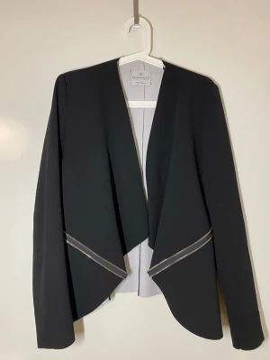 Svart kavaj från Rinascimento M - Snygg svart kavaj från Rinascimento med öppen front och coola dragkedjedetaljer vid höfterna. Jackan har lång ärm och en stilren, asymmetrisk skärning framtill. Perfekt för dig som gillar en modern och chic look.