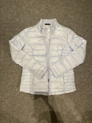 Ljusblå pufferjacka från Janina - Säljer en ljusblå pufferjacka från Janina i storlek 36. Jackan har quiltad design, dragkedja framtill och långärmade ärmar. Insidan är fodrad med ljusblått och vitt randigt tyg. Perfekt för kyliga dagar!