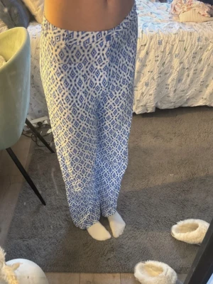 Blåvita mönstrade linnebyxor - Säljer ett par luftiga linnebyxor med blått och vitt grafiskt mönster. Byxorna har hög midja och rak passform, perfekta för en chill och avslappnad stil. Materialet känns lätt och svalt, vilket gör dem sköna att bära under varma dagar.