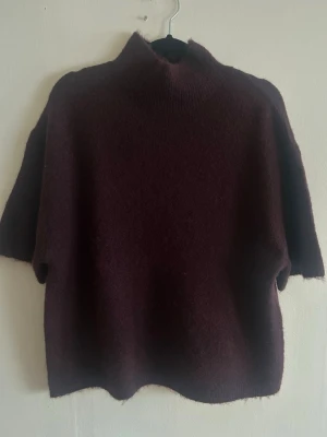 Vinröd stickad tröja Gina Tricot - Mysig vinröd stickad tröja från Gina Tricot i ullblandning. Tröjan har hög krage och korta ärmar, perfekt för lager på lager under höst och vinter. Enkel, oversized passform som ger en avslappnad vibe.