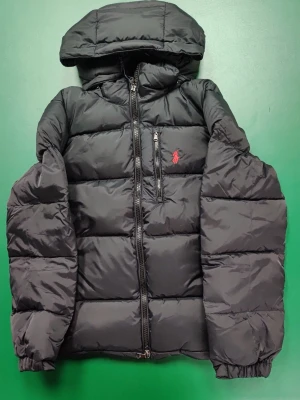 Svart pufferjacka från Polo Ralph Lauren - Säljer en svart pufferjacka från Polo Ralph Lauren med röd logga på bröstet. Jackan har huva, dragkedja framtill och en bröstficka med dragkedja. Perfekt för kalla dagar och riktigt skön att bära. Storlek S