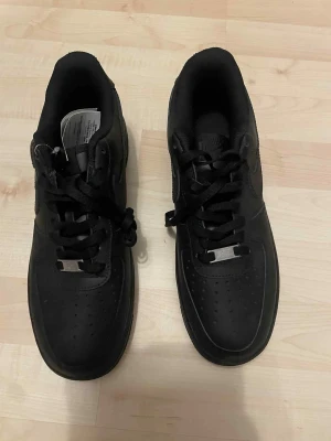 Nike Air Force 1 helsvarta sneakers - Säljer ett par klassiska Nike Air Force 1 i helsvart skinn. Skorna har rund tå, snörning och den ikoniska AF1-detaljen på snörena. Perfekt chunky sula och diskret Nike-logga på hälen. Grymma till alla outfits och riktigt clean look.
