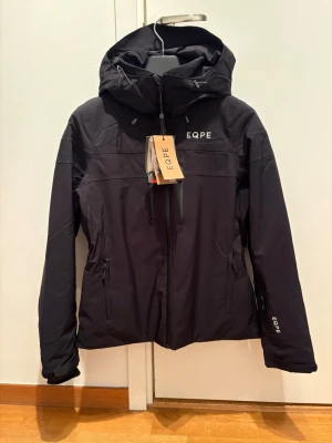 Svart skidjacka från EQPE dam S - Säljer en oanvänd svart skidjacka från EQPE i dammodell, storlek S. Jackan har huva med dragsko, dragkedja framtill och två sidofickor med dragkedja. Perfekt för dagar i backen. Enkel och stilren design med EQPE-logga på bröstet och ärmen. Hjälmkompatibel, fleecefodrad krage för extra komfort, ficka för liftkort. Vattenpelare: 10 000 mm. Andningsförmåga: 10 000g. RECCO-reflektorer. 