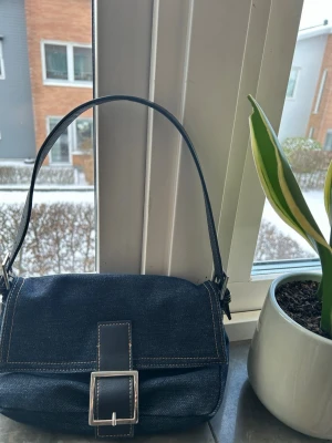 Mörkblå axelväska i denim - Snygg mörkblå axelväska i denim med bredt spänne framtill och kontrastsömmar. Väskan har ett klassiskt och stilrent uttryck med justerbart axelband och silverfärgat spänne. Perfekt för dig som gillar en trendig och enkel look.