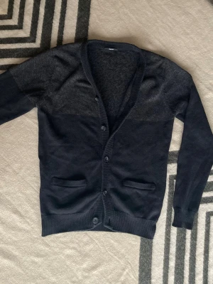 Mörkgrå slim fit kofta med knappar - Snygg mörkgrå kofta i slim fit-modell med ribbade muddar och knappstängning framtill. Koftan har två fickor och är tillverkad i ett stickat material som känns mjukt och bekvämt. Perfekt för dig som gillar stilrena och enkla plagg.