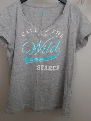 Grå t-shirt med tryck från Twintip - Snygg grå t-shirt från Twintip med vit och turkos text på bröstet. T-shirten har v-ringning och korta ärmar. Använd bara ett par gånger.