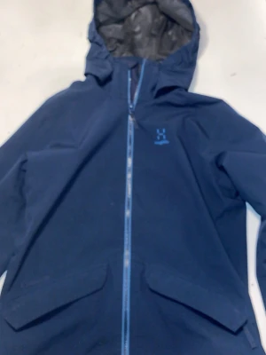 Blå vindjacka från Haglöfs - Säljer en blå vindjacka från Haglöfs med huva och hel dragkedja framtill. Jackan har två stora fickor med lock och reflekterande detaljer på ärmarna. Perfekt för dig som vill ha en stilren och funktionell jacka till vardags. Nästan ny skick, kom gjärna privat för bättre bilder och andra frågor