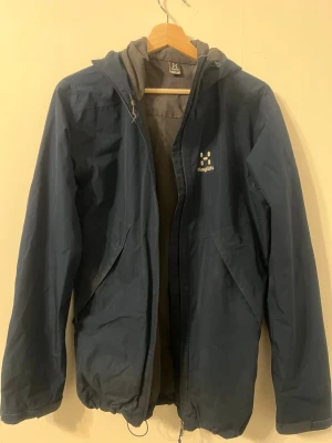 Blå vindjacka från Haglöfs - Snygg blå vindjacka från Haglöfs med huva och dragkedja framtill. Jackan har två stora fickor, justerbar nederkant och meshfoder på insidan. Perfekt för blåsiga dagar och outdoor-äventyr. Klassisk logga på bröstet och enkel, stilren design.