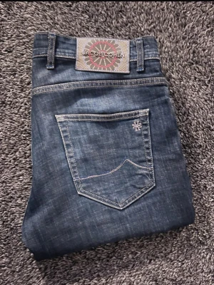 Nya Jacob Cohën jeans - Säljer ett par snygga blå jeans från Jacob Cohën i storlek 32. Klassisk femficksmodell med kontrastsömmar, broderad logga på bakfickan och coolt patch med mönster i midjan. Knapparna har JC-detaljer och insidan har ett unikt mönstrat tyg. Tillverkade i Italien.