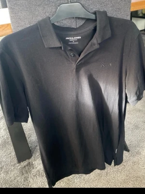 Svart pikétröja från Jack & Jones S - Klassisk svart pikétröja från Jack & Jones i storlek S. Tröjan har krage, två knappar framtill och diskret broderad logga på bröstet. Tillverkad i mjuk bomull som känns skön mot huden. Perfekt till jeans eller shorts för en clean look. Har aldrig använt den har bara legat i min gaderbob passar inte #mode #pike #Stilrent