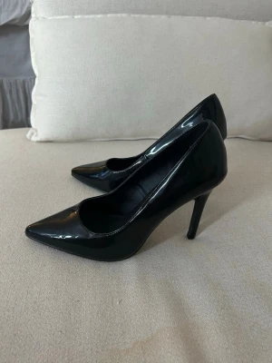 Svarta pumps med spetsig tå från Nelly - Säljer ett par klassiska svarta pumps från Nelly i glansigt syntetmaterial. Skorna har spetsig tå och hög klack som ger en snygg siluett. Perfekta för dig som vill ha en stilren och elegant look. 10 cm höga, även vi som är 38 i sko storlek kan ha dessa! 
