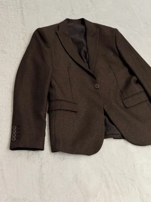 Mörkbrun kavaj från Daniel Gallotti - Snygg mörkbrun kavaj från Daniel Gallotti med klassisk passform. Kavajen har en knapp framtill, tre knappar vid ärmslutet och två fickor med lock. Fodret är svart och materialet känns mjukt och stilrent. Perfekt för dig som vill ha en clean och tidlös look.