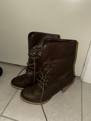 Bruna snörade boots med klack - Säljer ett par bruna boots i skinnimitation med snörning och robust klack. Skorna har rund tå, grov sula och metallfärgade öljetter. Perfekta för höst och vinter, ger en cool och stilren look till din outfit.
