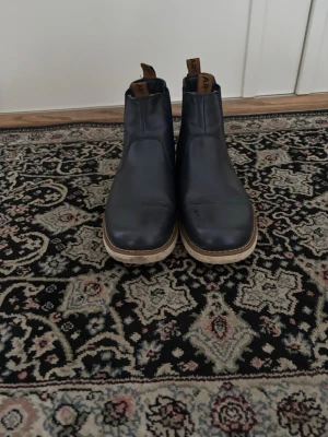 Mörkblå chelsea boots från Linear - Säljer ett par mörkblå chelsea boots från Linear i storlek 42. Skorna har elastiska sidopaneler, dragflik i brunt med logga och en rund tå. Utsidan är i skinn och insidan är fodrad med mjukt material för extra komfort. Sulan är ljus och platt med kontrastsöm.