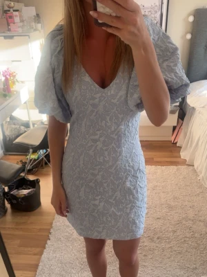 Ljusblå kortklänning med puffärm - Supersöt ljusblå kortklänning med v-ringning och stora puffärmar. Klänningen har ett diskret blommigt mönster och figurnära passform som ger en riktigt fin siluett. Perfekt för sommarens alla tillfällen! Endast använd en gång🥰