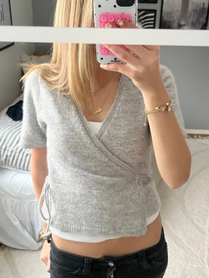 Grå omlottkofta från Gina Tricot - Mysig ljusgrå omlottkofta från Gina Tricot i ullblandning. Kortare modell med knyt i sidan och v-ringad hals. Perfekt till lager på lager och funkar året runt. Kort ärm och mjuk stickad känsla.