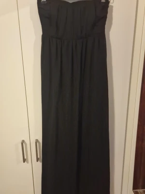 Svart axelbandslös långklänning - Säljer en svart axelbandslös långklänning med smockad överdel och lätt utställd kjol. Klänningen har en enkel och elegant look med mjukt fallande tyg, perfekt för dig som gillar stilrena plagg. Materialet känns lätt och flowy. Det finns band med om man vill ha det. 