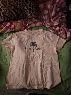 Beige Côte d'Azur t-shirt med tryck - Snygg beige t-shirt med grönt palmtryck och texten 'Côte d'Azur France' på bröstet. Klassisk rund hals och korta ärmar. Perfekt för dig som gillar chill och enkel stil med en hint av semesterkänsla.