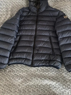 Svart pufferjacka från Colmar - Säljer en mörkblå pufferjacka från Colmar med klassisk quiltad design och hög krage. Jackan har två sidofickor med dragkedja och Colmar-logga på ärmen. Perfekt för kalla dagar och riktigt snygg till alla outfits.