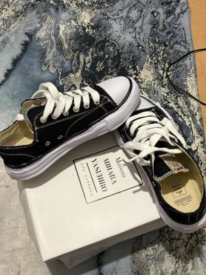 Maison Mihara Yasuhiro svarta sneakers - Maison Mihara Yasuhiro sneakers i svart canvas med vita kontrastsömmar och tjock vit sula. Skorna har rund tå, vita snören och en unik, vågig sula som ger en cool twist. Klassisk low-top modell med retrovibbar och tydlig streetwear-känsla.