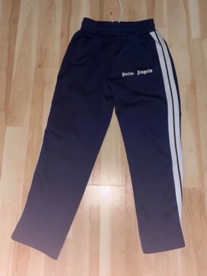 Palm Angels blå träningsbyxor S - Säljer ett par mörkblå träningsbyxor från Palm Angels i storlek S. Byxorna har vita sidoränder, resår i midjan och dragkedjor vid bensluten. Tillverkade i 100% polyester för en sportig vibe och bekväm passform. Perfekta för dig som gillar streetwear.