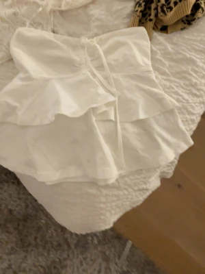 Vit volang topp med snörning - Supersöt vit topp med dubbla volanger och snörning framtill. Modellen är ärmlös och croppad, perfekt till jeansshorts eller kjol. Tunt och lätt material som känns luftigt under varma dagar.