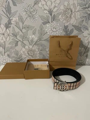 Rutig Burberry bälte med silverdetalj - Säljer ett klassiskt Burberry bälte med ikoniskt rutigt mönster i beige, svart, rött och vitt. Bältet har ett stort, runt spänne i silver med Burberrys logotyp. Kommer med originalpåse och ask. Perfekt accessoar för att lyfta vilken outfit som helst.