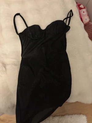 Glittrig svart kort klänning - Supersnygg svart kort klänning med tunna axelband och glittrigt tyg. Klänningen har en figurnära passform och markerad byst, perfekt för att sticka ut. Materialet känns mjukt och stretchigt, vilket gör den bekväm att bära hela kvällen.