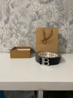 Rutig Burberry bälte med B-spänne - Säljer ett klassiskt Burberry bälte med ikoniskt rutigt mönster i beige, svart och rött. Bältet har ett stort silvrigt B-spänne framtill och insidan är fodrad med Burberrys signaturrutor. Kommer med originalpåse och box.