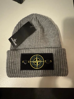 Grå ribbstickad mössa Stone Island - Säljer en grå ribbstickad mössa från Stone Island med den klassiska logotypen framtill på en svart patch. Topp skick, skönt till de kalla dagarna 