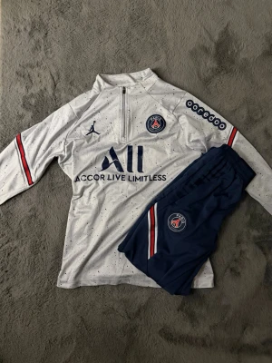 PSG träningsset - PSG träningsset från Jordan Brand med vit långärmad tröja (halv zip) och mörkblå byxor. PSG- och Jordan-loggor samt sponsortryck. Sportig passform, perfekt för träning eller vardag.