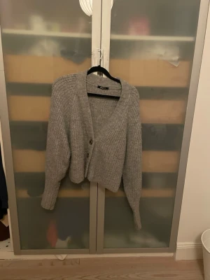 Grå stickad kofta från Gina Tricot - Mysig grå stickad kofta från Gina Tricot med v-ringning och två knappar framtill. Koftan har lång ärm och en loose passform som gör den perfekt att slänga på sig under kyliga dagar.