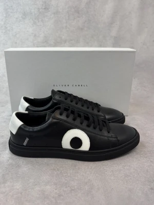 Svarta sneakers från Oliver Cabell - Snygga svarta sneakers från Oliver Cabell i skinn med vita detaljer på hälen och en stor vit cirkel på sidan. Klassisk låg modell med svart snörning och platt sula. Perfekta för dig som gillar stilrena och moderna skor med en twist. Box och dustbag medföljer vid köp! Pris kan diskuteras 