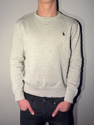 Ljusgrå sweatshirt från Polo Ralph Lauren - Säljer en stilren ljusgrå sweatshirt från Polo Ralph Lauren med klassisk rund halsringning och broderad marinblå logga på bröstet. Tröjan har normal passform och är tillverkad i mjuk bomull, perfekt för både chill och snygga outfits. Utmärkt skick. Hör av er för fler frågor/bilder eller funderingar😊Tar emot prisförslag och direkt köp