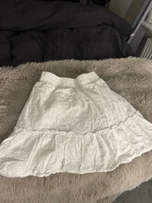 Vit kortkjol med volang och broderi - Supersöt vit kortkjol med volangkant och broderade blommor. Kjolen har resår i midjan och är gjord i ett lätt bomullstyg som känns perfekt för varma dagar. Den romantiska stilen gör den enkel att matcha med både toppar och sneakers. Storlek XS men passar mig som är S.