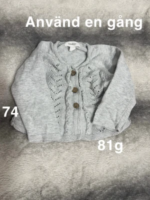 Ljusgrå stickad kofta från Newbie - Supersöt ljusgrå stickad kofta från Newbie med fina träknappar och dekorativt hålmönster framtill. Perfekt till lager på lager och enkel att matcha med andra plagg. Långärmad och mjuk i materialet.🌸 Använd en gång. Storlek 74.