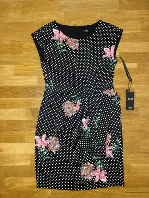 Svart prickig klänning s.Oliver - Supersnygg svart kortklänning från s.Oliver med vita prickar och färgglada blommor i rosa och orange. Klänningen har en figurnära passform, är ärmlös och har dragkedja bak. Perfekt för dig som vill sticka ut med både mönster och färg!