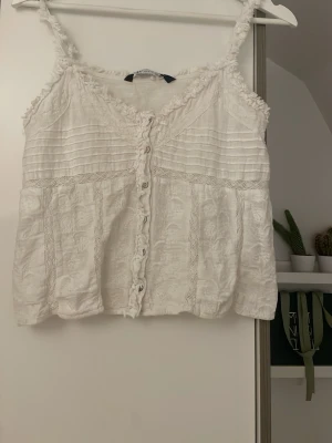 Vit broderad linnetopp från Zara - Supersöt vit linnetopp från Zara med broderade detaljer och spetsinslag. Toppen har smala axelband, knappar framtill och volangkant längs ringningen. Perfekt för varma dagar och ger en romantisk vibe till din outfit.storlek S, hör av dig om du har några frågor eller funderingar 💕