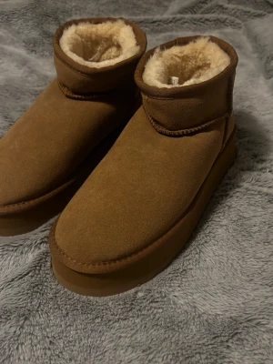 Bruna boots i mocka - Säljer ett par klassiska bruna boots med mjukt fårskinnsfoder och rund tå, inspirerade av ett liknande märke. Skorna har en platt sula och är platform. Perfekta för kalla dagar och ger en cozy vibe.