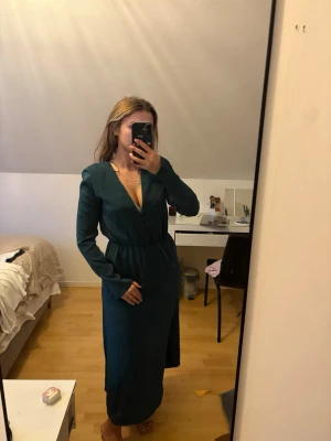 Mörkgrön långklänning - Säljer en stilren mörkgrön långklänning från DW Ella i storlek S. Klänningen har v-ringning, långa ärmar och markerad midja med resår. Tyget är mjukt och faller snyggt längs kroppen. Perfekt för dig som gillar en enkel men elegant look.aldrig använd har bara stått o dammat I garderoben prislappen e kvar! Nypris 700kr! 