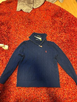 Marinblå kabelstickad tröja Polo Ralph Lauren - Kabelstickad marinblå tröja från Polo Ralph Lauren med halv dragkedja och hög krage. Tröjan har lång ärm, ribbade muddar och en röd broderad logga på bröstet. Perfekt för lager-på-lager och snygg till jeans eller chinos.